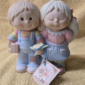 May include: A porcelain figurine of a boy and girl holding hands. The boy is wearing blue overalls and a rainbow striped shirt. The girl is wearing a pink shirt and a blue skirt. They are both smiling. The figurine is holding a small white flower.
