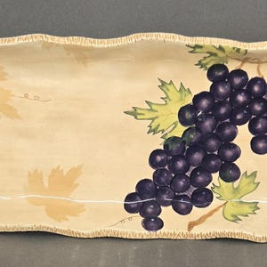May include: A rectangular ceramic serving tray with a cream-colored base and a textured, scalloped edge. The tray features a painted illustration of a large cluster of dark purple grapes with green leaves on the right side and faint leaf silhouettes.