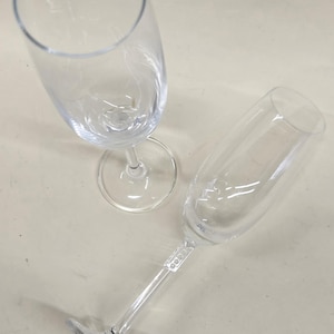 May include: Two clear champagne flutes on a light beige surface. One flute is upright, and the other is lying on its side. The flutes have a long stem and a wide bowl, perfect for celebratory drinks.