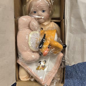 May include: A collectible doll with blue eyes and a yellow bonnet, packaged in a cardboard box. The box also contains a pink plush toy, a Pluto figurine, and a Pluto-themed handkerchief.