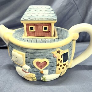 May include: A ceramic teapot shaped like Noah's Ark, featuring a small house on top and animal figures on the sides. The teapot is painted in pastel colors, including blue, yellow, and brown, with a heart-shaped detail.