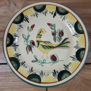 May include: A white ceramic plate with a yellow bird and green foliage design. The plate has a green and yellow border with a scalloped edge.