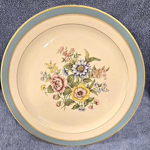 May include: A round, cream-colored plate with a light blue border and gold trim. The plate features a floral bouquet in the center with pink, yellow, blue, and white flowers. A classic design for serving food.