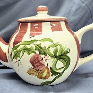 May include: Cream-colored ceramic teapot with a lid, handle, and spout. The teapot features red and white striped accents and is decorated with green leaves, red fruit, and brown pinecones. The teapot is designed for brewing tea.
