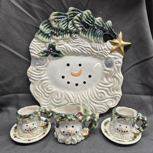 May include: A ceramic snowman-themed serving set. The set includes a large plate with a snowman face and a Christmas tree-like border, a creamer, and two teacups with saucers. The snowman has a carrot nose and black dot eyes.