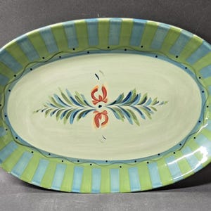 May include: An oval ceramic serving platter featuring a light green center with a central painted floral motif in orange, blue, and green. The rim is decorated with alternating vertical stripes of light green and blue, accented by a thin wavy line.