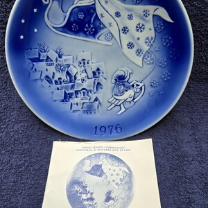 May include: A blue and white decorative plate from 1976, featuring a winter scene with a sleigh and snowflakes. The plate has a detailed illustration of a town and a figure in a sleigh. Includes a paper with the same design.