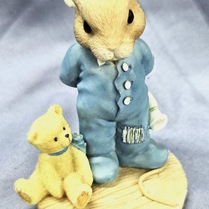 May include: A figurine of a rabbit wearing a blue jumpsuit with three white buttons, standing on a wooden base. A yellow teddy bear with a blue bow sits beside the rabbit. The figurine has a whimsical, storybook aesthetic.