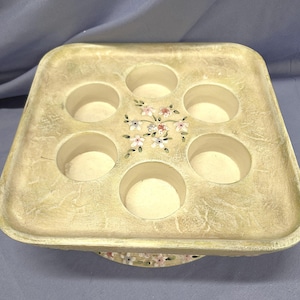 May include: A square, off-white decorative tray with six circular indentations. The tray features a floral design with pink and white flowers. The tray has a distressed, textured finish, and is likely made of ceramic or a similar material.