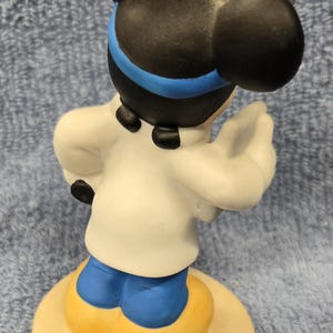 May include: A ceramic figurine of a cartoon mouse character, viewed from the back. The mouse has black ears, a white coat, blue pants, and yellow shoes. The figurine is standing on a round, beige base. The mouse has a blue headband.