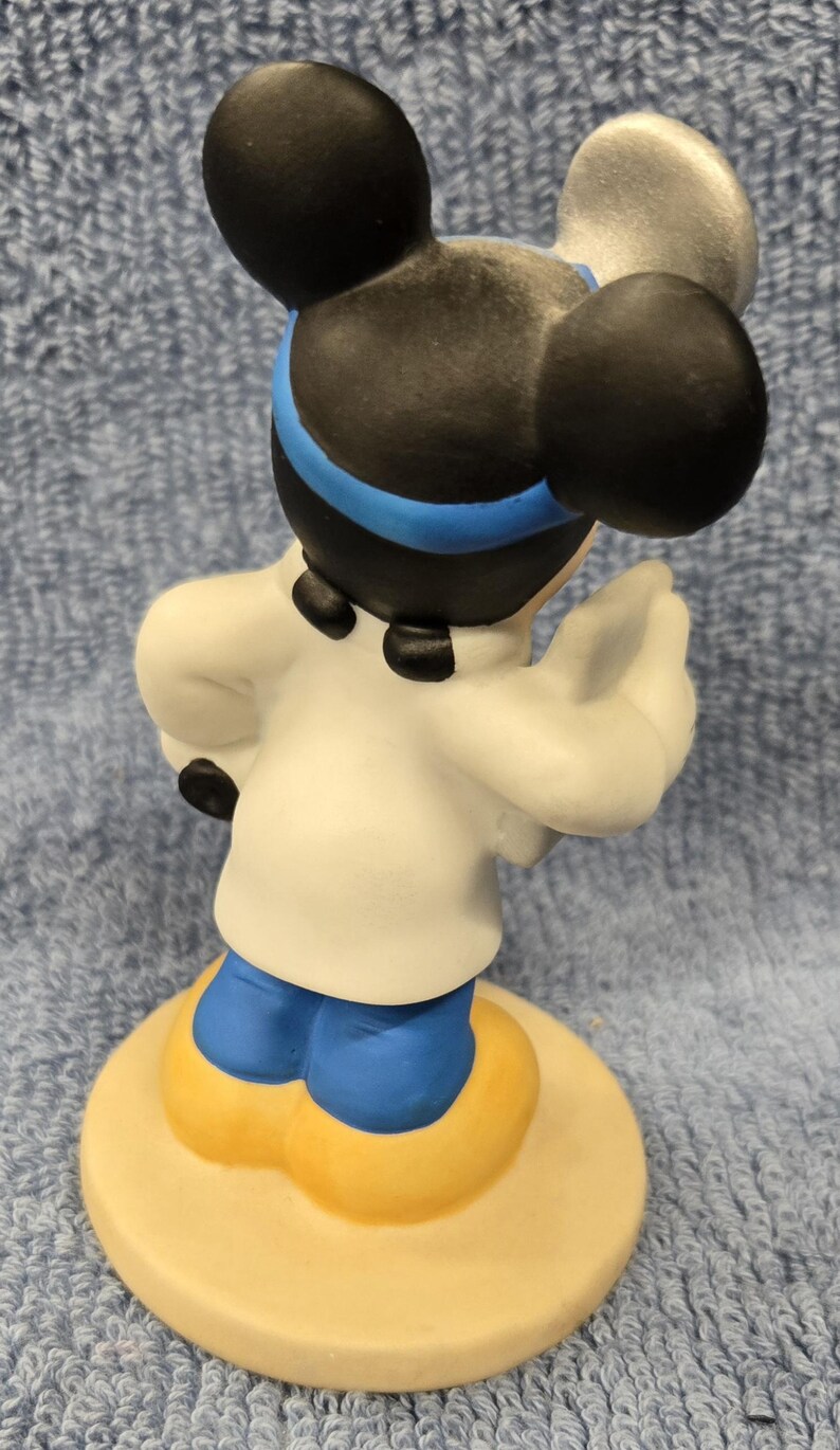 May include: A ceramic figurine of a cartoon mouse character, viewed from the back. The mouse has black ears, a white coat, blue pants, and yellow shoes. The figurine is standing on a round, beige base. The mouse has a blue headband.