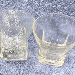 May include: Two clear, textured glass tumblers. The glasses have a unique, organic shape with a slightly flared rim and a textured surface. The glasses are empty and appear to be made of thick glass.