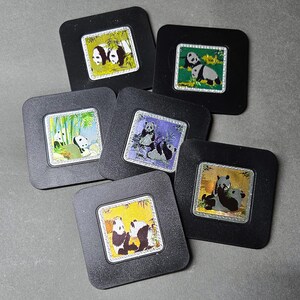May include: A set of six square black coasters, each featuring a central inset illustration of giant pandas in various natural settings. The illustrations depict pandas interacting with bamboo, rocks, and foliage, framed by a silver-toned border.