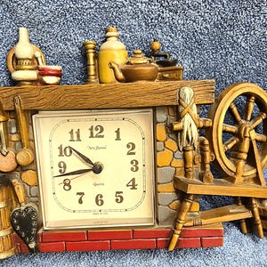 May include: A decorative quartz clock with a rustic theme. The clock features a square face with black numerals and hands, set within a fireplace mantel design. The mantel includes miniature kitchen tools, a spinning wheel, and a barrel.