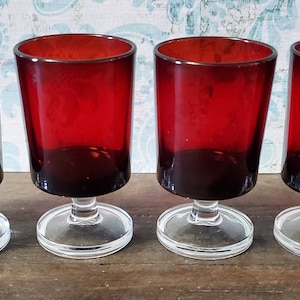 May include: Four red glass goblets with clear glass stems and bases. The goblets are arranged in a row on a wooden surface.