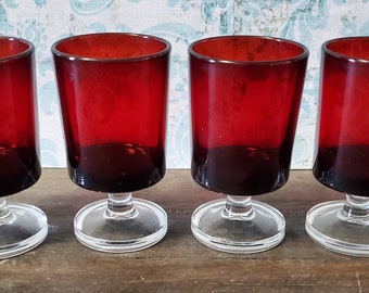 May include: Four red glass goblets with clear glass stems and bases. The goblets are arranged in a row on a wooden surface.