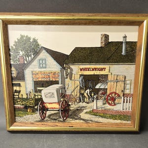 May include: A framed print depicting a wheelwright shop with a horse-drawn carriage in the foreground. The scene features a building with the sign WHEELWRIGHT, a person working inside, and red-wheeled carriages. The frame is wood with gold trim.