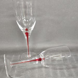 May include: Two clear glass wine glasses with long stems. Each stem features a distinct, elongated red glass accent that extends from the base of the bowl down toward the circular foot, creating a striking contrast against the clear glass.