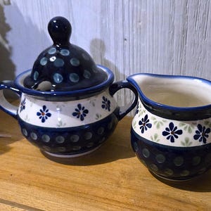 May include: A ceramic sugar bowl with a lid and a creamer. Both pieces are decorated with a blue and white floral pattern and a dark blue polka dot design. The sugar bowl has a handle and the creamer has a spout.