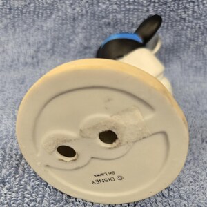 May include: A ceramic figurine of a cartoon character, featuring a black head with blue accents and white body. The base is white with a light tan rim. The bottom of the base has two holes and the text "&copy; DISNEY Sri Lanka".