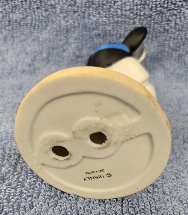 May include: A ceramic figurine of a cartoon character, featuring a black head with blue accents and white body. The base is white with a light tan rim. The bottom of the base has two holes and the text "&copy; DISNEY Sri Lanka".