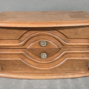 May include: A rectangular wooden jewelry box with a medium brown finish. The front features two stacked drawers, each with a circular, textured metal knob. The wood grain is visible, and the design includes carved, concentric, eye-shaped patterns.