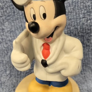 May include: A ceramic figurine of Mickey Mouse dressed as a doctor. Mickey wears a white lab coat, a red tie, and a blue headband. He has yellow shoes and a silver medical mirror on his head. The figurine stands on a beige base.