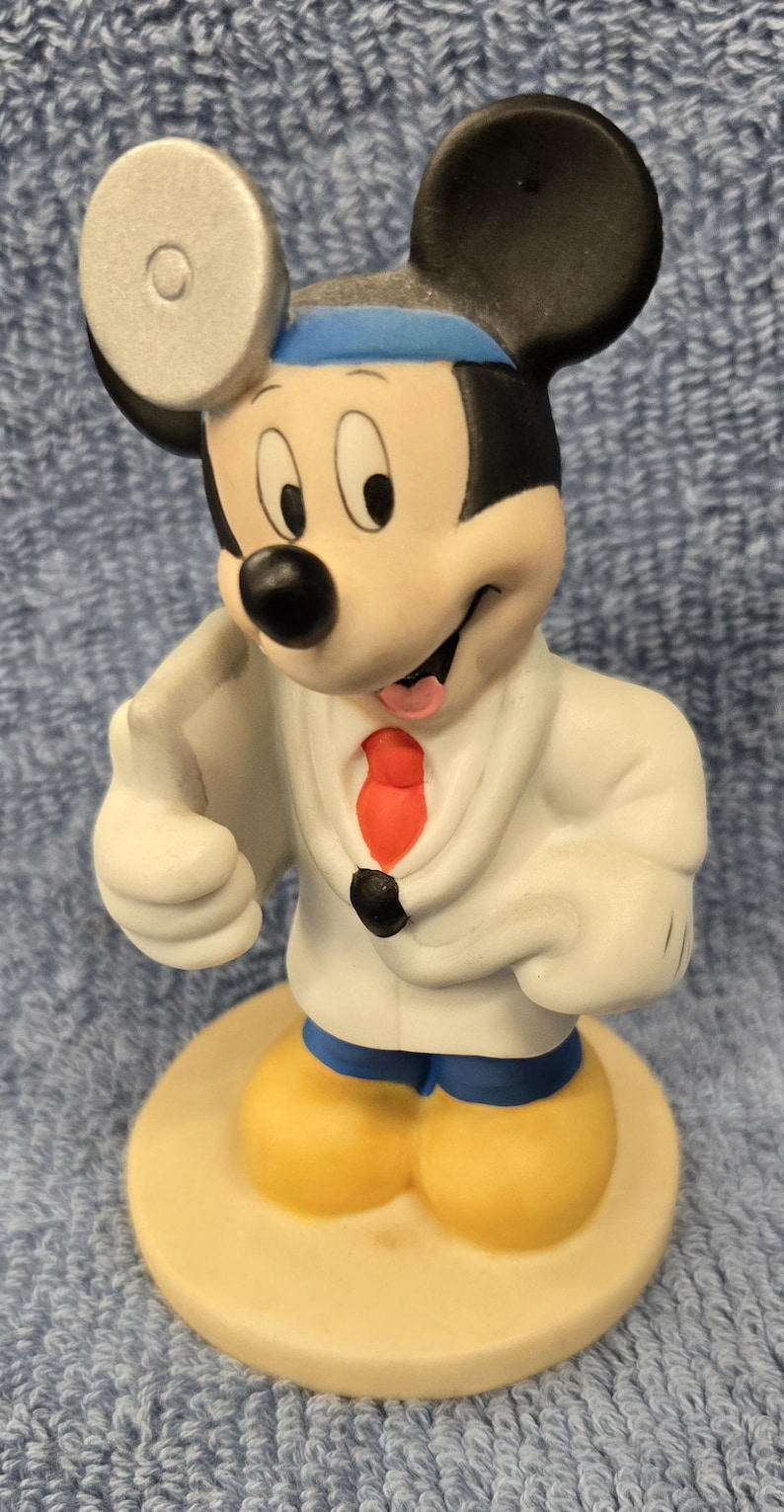 May include: A ceramic figurine of Mickey Mouse dressed as a doctor. Mickey wears a white lab coat, a red tie, and a blue headband. He has yellow shoes and a silver medical mirror on his head. The figurine stands on a beige base.