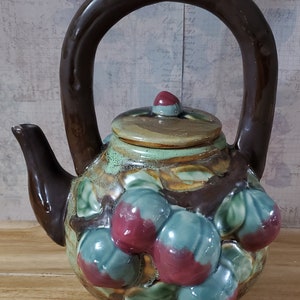 May include: A ceramic teapot with a large brown handle and spout. The body is decorated with raised, colorful fruit and leaves. The lid has a small red knob. The teapot is a unique piece of kitchenware.