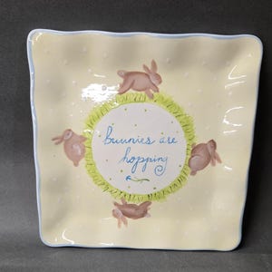 May include: A square, pale yellow ceramic plate with a wavy blue rim and white polka dots. The center features a circular green grassy patch with four brown rabbits surrounding the text "bunnies are hopping" written in blue cursive script.