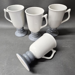 May include: A set of four ceramic mugs featuring a white cylindrical body and a glossy grey pedestal base. Each mug has a curved handle on the side. Three mugs are standing upright, while one mug is positioned lying on its side.
