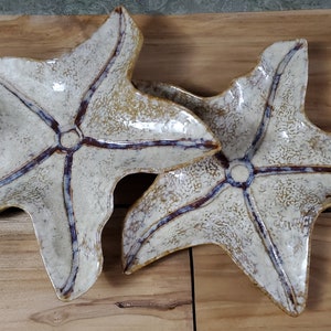 May include: Two ceramic starfish-shaped dishes with a white glaze and brown and blue accents. The dishes are on a wooden surface.