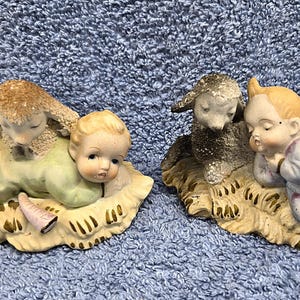 May include: Two ceramic figurines depicting a baby and a lamb resting on a bed of hay. The baby is wearing a light green outfit, and the lamb has a fluffy, textured coat. The second figurine shows a baby in a blue outfit and a gray lamb.
