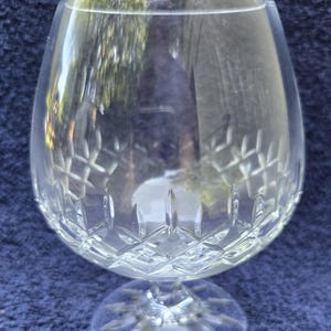 May include: A clear, stemmed brandy snifter with a wide bowl and a patterned design near the base. The glass reflects the surrounding environment. The snifter sits on a blue surface.