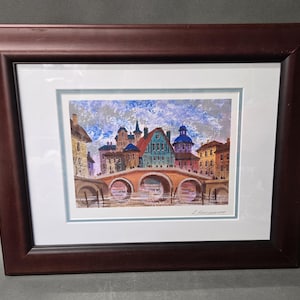 May include: A framed painting depicting a cityscape with a stone bridge over a river. The scene features colorful buildings with varied rooflines under a textured blue and grey sky. The artwork is enclosed in a dark brown wooden frame with a white mat.
