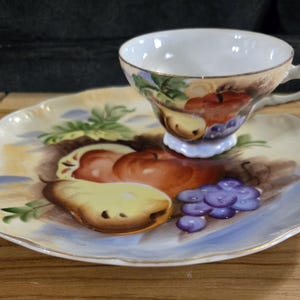 May include: A white porcelain teacup and saucer set with a hand-painted design of fruit, including pears, apples, and grapes. The set features a light blue and yellow background with gold accents.