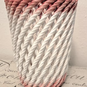 May include: A white ceramic vase with a woven rope design. The top and bottom edges are painted with a gradient of pink. The vase has a cylindrical shape and is suitable for home decor.