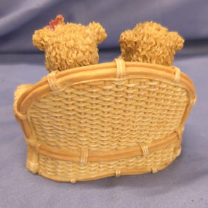 May include: A decorative figurine of two teddy bear figures seated on a woven wicker-style bench. The bears have textured fur and are light brown. The bench is a light tan colour.