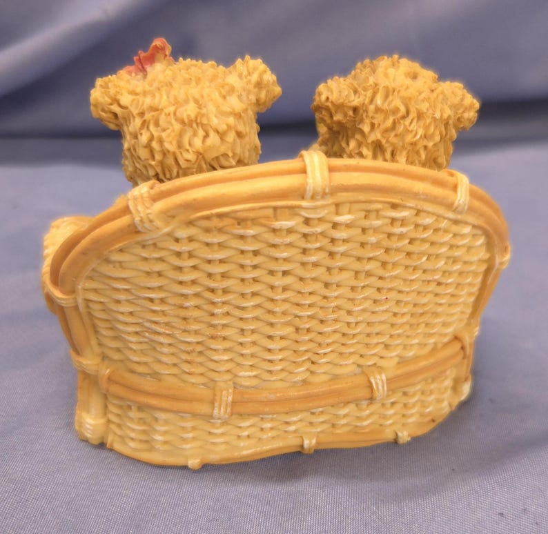May include: A decorative figurine of two teddy bear figures seated on a woven wicker-style bench. The bears have textured fur and are light brown. The bench is a light tan colour.