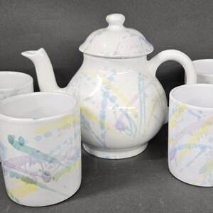 May include: A white ceramic tea set consisting of one teapot with a lid and four matching mugs. Each piece features a decorative paint splatter pattern in pastel shades of yellow, light blue, and soft purple across the smooth white surface.