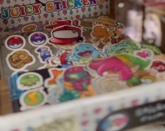 Op de afbeelding: Een transparante plastic doos gevuld met kleurrijke stickers met cartoonfiguren. De doos heeft een roze en blauw hartlogo met de tekst "Juicy Stickers".