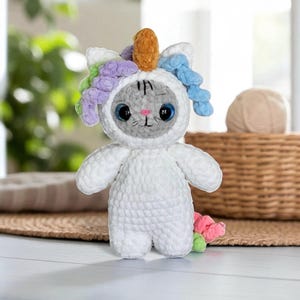 Puede incluir: Un gato de ganchillo blanco hecho a mano disfrazado de unicornio. El peluche tiene una cara gris con ojos azules grandes, una nariz rosa y una pequeña sonrisa. El unicornio presenta un cuerno dorado y pelo de hilo colorido.