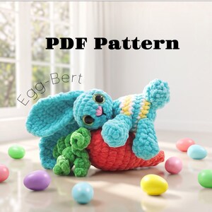 May include: A crochet PDF pattern titled Egg-Bert featuring a plush turquoise bunny with large dark eyes and a pink nose, resting on a large textured red carrot with green leafy tops, surrounded by small colorful plastic Easter eggs.