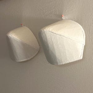 May include: Two off-white, curved, padded shoulder inserts featuring visible stitched edges. Each piece is secured to a textured surface with a single small pin topped with a pink bead, highlighting the structured, molded shape of the garment components.