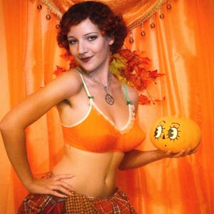 May include: An orange bralette with white trim and green straps, paired with a plaid skirt. A silver pentagram necklace is worn. The item is held alongside a small orange pumpkin featuring a hand-drawn cartoon face with large eyes and a nose.