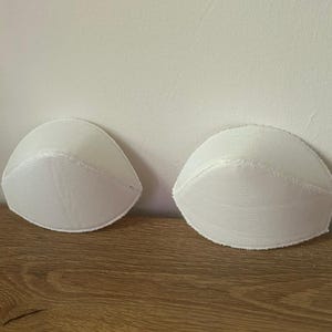 May include: A pair of white, molded bra cups with a smooth, rounded shape and stitched edges. These semi-circular inserts are designed for garment construction, featuring a clean, solid white fabric surface and a structured, curved profile.