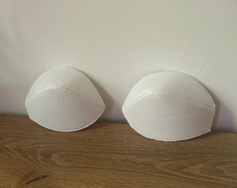 May include: A pair of white, moulded bra cups with a smooth, rounded shape and stitched edges. These foam inserts are designed for garment construction and provide structure, featuring a clean, minimalist appearance with a consistent texture.