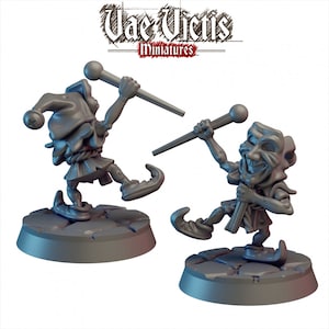 May include: Two grey painted miniature figurines of jesters holding staffs. The jesters are wearing jester hats and costumes. The figurines are standing on round bases.