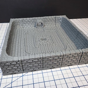 May include: A gray, square miniature dungeon terrain piece with a brick-like texture. The interior features a recessed area with a circular drain and a cobblestone pattern. The walls are also made of brick-like material.