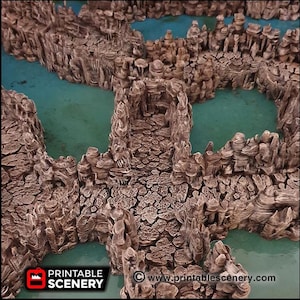 Puede incluir: Una vista cercana de un modelo de terreno impreso en 3D para juegos de mesa. El modelo presenta un paisaje rocoso parecido a un cañón con un camino que atraviesa el centro. El camino está rodeado de formaciones rocosas altas y dentadas y una pequeña piscina de agua.